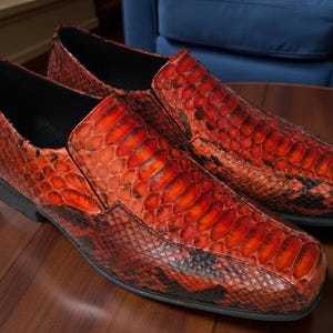 Könnte beinhalten: Ein Paar rot-schwarz gemusterte Slipper im Schlangenleder-Look. Die Schuhe haben einen niedrigen Absatz und eine runde Spitze. Das Leder hat ein glänzendes Finish, und die Schuhe werden auf einer Holzoberfläche präsentiert.