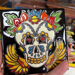 May include: Square black ceramic tile featuring a colorful sugar skull design. The skull has a white base with intricate details, including heart-shaped eyes and floral accents in red, blue, yellow, and orange. The skull has yellow and orange wings.