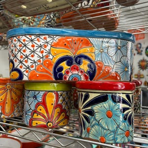 Puede incluir: Una maceta rectangular de cerámica con borde azul y un patrón floral pintado a mano en naranja, amarillo y azul oscuro. Debajo, dos macetas cilíndricas de cerámica presentan diseños coloridos similares con bordes rojo y verde.