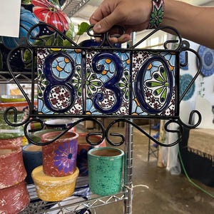 Może przedstawiać: Dekoracyjna tablica adresowa z numerami "830" na trzech ceramicznych płytkach. Płytki mają niebiesko-biały kwiatowy wzór, osadzone w czarnej ramie z kutego żelaza. Inne kolorowe ceramiczne doniczki znajdują się na półce poniżej.