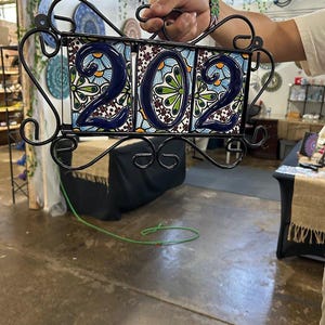 Pode incluir: Placa de endereço decorativa com o número "202" em um vibrante design floral de azulejos azuis, verdes e brancos. Os azulejos estão inseridos em uma moldura de ferro forjado preto com volutas decorativas. Um presente de boas-vindas único.