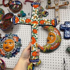 May include: A hand-painted ceramic cross featuring a white base decorated with vibrant orange flowers and green leaves. The cross has blue and orange accents at the ends and a small twine loop at the top for hanging, showcasing a glossy finish.