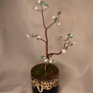 May include: A handcrafted decorative tree sculpture. The tree features a copper wire trunk and branches adorned with small, colorful gemstones. It is planted in a small, dark brown ceramic pot with a decorative rim, filled with moss.
