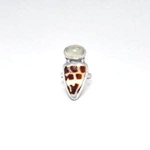 May include: A silver ring featuring a teardrop-shaped shell inlay with a brown and white spotted pattern, topped with a smooth, translucent, pale circular stone. The metal setting has a polished finish and holds both elements securely in place.