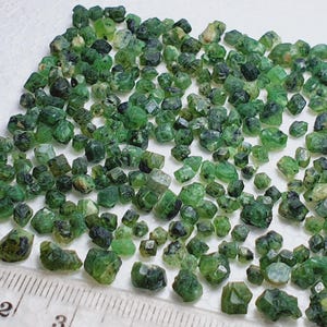 Könnte beinhalten: Eine Ansammlung kleiner, roher grüner Edelsteine unterschiedlicher Größe, die auf einer weißen Oberfläche verstreut sind. Die Edelsteine weisen eine kristalline Struktur und eine Reihe von Grüntönen auf. Ein Lineal mit Zentimetermessungen liegt zur Größenbestimmung bereit.