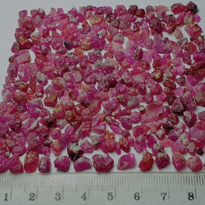 Könnte beinhalten: Eine Sammlung kleiner, grob geschnittener, rosa Edelsteine. Die Edelsteine sind auf einer weißen Oberfläche verstreut, mit einem Lineal am unteren Rand. Die Edelsteine variieren in Größe und Farbton, wobei einige röter erscheinen als rosa.