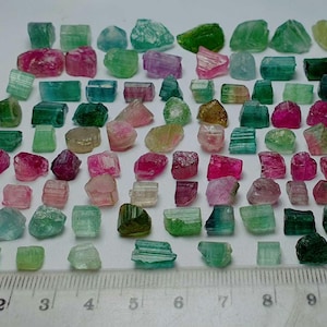 May include: A collection of raw tourmaline gemstones in various colors, including pink, green, and blue. The gemstones are irregularly shaped and arranged on a white surface next to a ruler showing inches.
