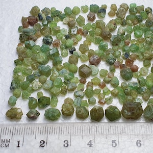 May include: A collection of small, faceted green gemstones, with hints of brown and yellow, are scattered across a white surface. A ruler with centimeter and millimeter markings is at the bottom, indicating size. The gemstones vary in size and shape.