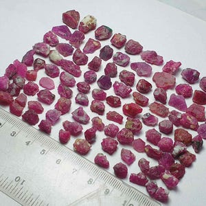 May include: A collection of raw ruby gemstones in shades of red and pink, with some showing white and green inclusions. A ruler with centimetre markings is placed next to the gemstones.