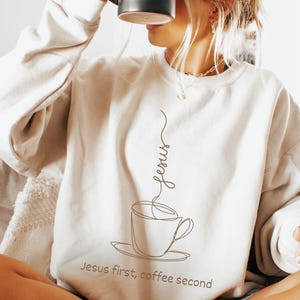 Könnte beinhalten: Cremefarbenes Sweatshirt mit Kaffeetassen-Design und dem Text "Jesus first, coffee second." Eine Person hält eine schwarze Kaffeetasse. Das Sweatshirt hat einen Rundhalsausschnitt und lange Ärmel. Das Design ist minimalistisch.