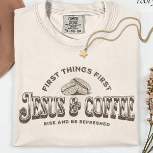 Puede incluir: Camiseta de color marfil con el texto "FIRST THINGS FIRST JESUS & COFFEE RISE AND BE REFRESHED" en marrón. La camiseta presenta un gráfico de granos de café. Un collar dorado con un colgante de estrella descansa sobre la camiseta.
