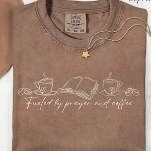 Op de afbeelding: Espresso-kleurig T-shirt met de witte tekst "Fueled by prayer and coffee". Het ontwerp bevat lijntekeningen van koffiekopjes, koffiebonen en een open boek. Een gouden sterhangerketting ligt op het shirt.