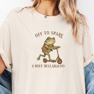 以下が含まれることがあります： アイボリー色のTシャツ。スクーターに乗ったカエルの漫画が描かれています。カエルはベストとスカーフを着ています。「OFF TO SPARK A HOLY HULLABALOO」というテキストが茶色でプリントされています。Tシャツの袖はロールアップされています。