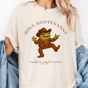 Puede incluir: Camiseta color marfil con una rana de dibujos animados vestida con un atuendo de vaquero. El texto "HOLY HOOTENANNY" está sobre la rana, con "make a joyful noise" debajo. La rana lleva sombrero, chaqueta y botas marrones.