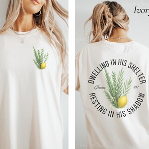 May include: Ivory-colored t-shirt with a botanical design featuring green leaves and a yellow lemon on the front. The back of the shirt has a circular design with text and the same botanical illustration. The text reads "Dwelling in His Shelter, Resting in His Shadow, Psalm 91:4."