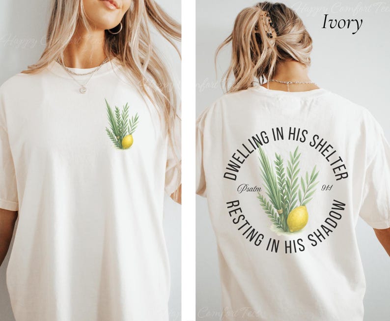 May include: Ivory-colored t-shirt with a botanical design featuring green leaves and a yellow lemon on the front. The back of the shirt has a circular design with text and the same botanical illustration. The text reads "Dwelling in His Shelter, Resting in His Shadow, Psalm 91:4."