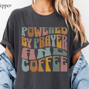 May include: Dark grey t-shirt featuring the text "POWERED BY PRAYER AND COFFEE" in a retro, multi-coloured design. The shirt has rolled-up sleeves and a relaxed fit. The word "Pepper" is in the top left corner.