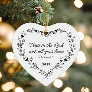 May include: A white heart-shaped Christmas ornament with a gold string for hanging. The ornament features the text "Trust in the Lord with all your heart" and "Proverbs 3:5" in black script, surrounded by a black floral design. The year "2025" is also displayed.