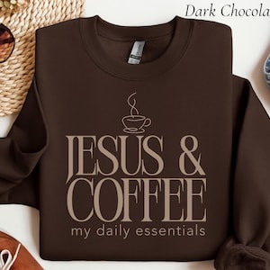 May include: A dark chocolate brown crewneck sweatshirt featuring a graphic design in light beige. The design includes a line-art coffee cup icon above the text "JESUS & COFFEE" in large serif letters, followed by "my daily essentials" in smaller text.