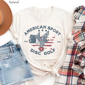 以下が含まれることがあります： クリーム色のTシャツ。ワシ、アメリカ国旗、そして「AMERICAN SPORT DISC GOLF」の文字と「EST. 1976」の文字が描かれた、かすれたグラフィックが特徴です。このシャツは、デニムショーツとチェック柄のシャツと組み合わせています。