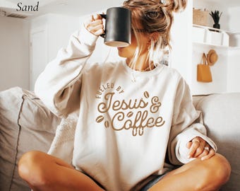May include: A sand-colored sweatshirt with the text "Fueled by Jesus & Coffee" in brown. The sweatshirt features a crew neck and long sleeves. A black mug is held up to the face. The overall theme is faith and coffee.