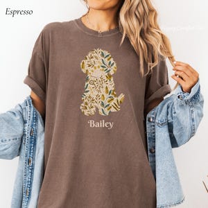 May include: Espresso-colored t-shirt featuring a floral dog silhouette with the name "Bailey" below. The dog design incorporates green, yellow, and white botanical elements. The shirt has rolled-up sleeves and a relaxed fit.