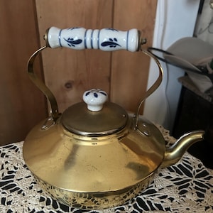 May include: A vintage brass teapot with a white and blue floral patterned handle and lid knob. The teapot has a rounded body, a spout, and a curved handle. The teapot is sitting on a cream-colored, open-weave doily.