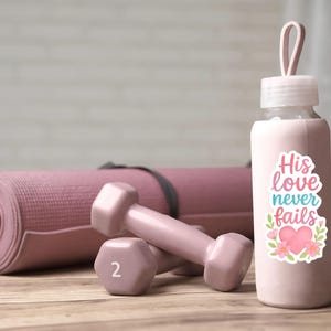 May include: A pink water bottle with a sticker that reads "His love never fails" next to two pink 2-pound dumbbells and a rolled-up yoga mat. The items are on a wooden surface.