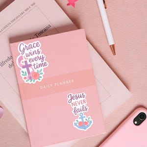 May include: A pink daily planner with the text "Grace wins every time" and "Jesus never fails" on stickers. A white pen and a pink smartphone are also visible. The planner is on a pink surface.