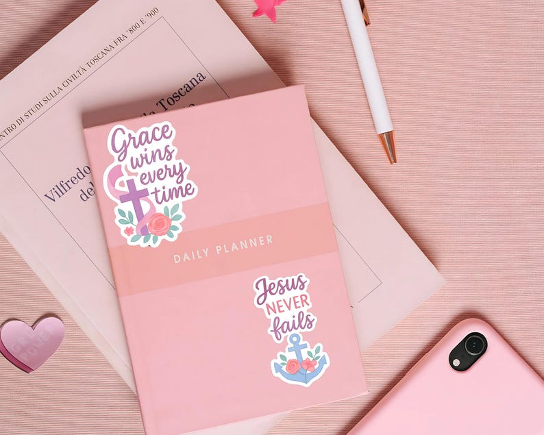 May include: A pink daily planner with the text "Grace wins every time" and "Jesus never fails" on stickers. A white pen and a pink smartphone are also visible. The planner is on a pink surface.