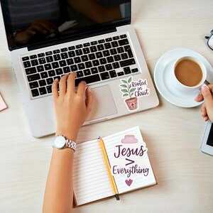 May include: A laptop with a sticker that says "Rooted in Christ" next to a coffee cup and a notebook that says "Jesus > Everything". A person is using the laptop and holding the coffee cup. Other items include a tablet, glasses, and a watch.