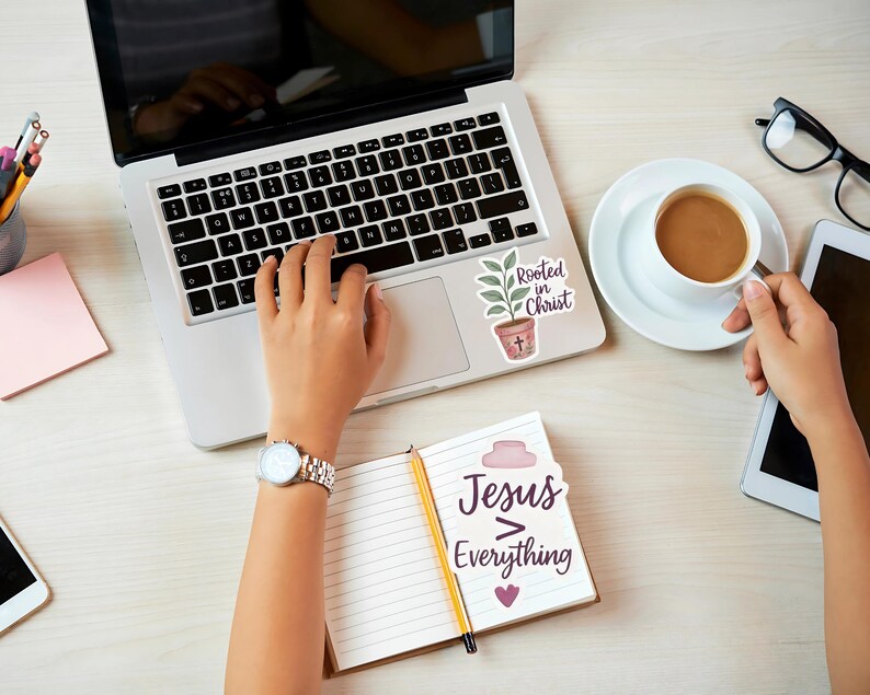 May include: A laptop with a sticker that says "Rooted in Christ" next to a coffee cup and a notebook that says "Jesus > Everything". A person is using the laptop and holding the coffee cup. Other items include a tablet, glasses, and a watch.