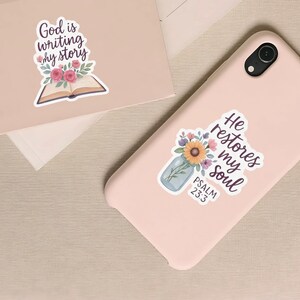 May include: Two stickers with religious quotes and floral designs. One sticker features the text "God is writing my story" above an open book with flowers. The other sticker says "He restores my soul" with a floral arrangement and the text "Psalm 23:3".