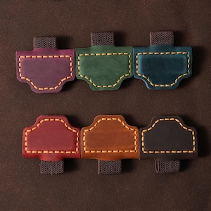 Puede incluir: Seis pestañas de diario de cuero en varios colores: morado, verde, turquesa, rojo, marrón y negro. Cada pestaña tiene una costura en contraste y un lazo elástico oscuro para sujetar. También se ve un marcapáginas de hoja metálica.