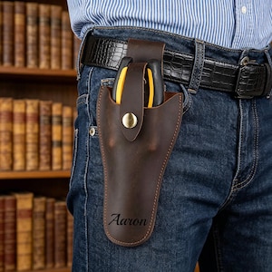 Puede incluir: Una funda de cuero marrón oscuro para herramientas con cierre de broche y el nombre Aaron grabado en la parte frontal. La funda está sujeta a un cinturón negro texturizado sobre pantalones vaqueros azules, conteniendo una herramienta con mangos amarillos y negros.