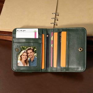 Puede incluir: Cartera de cuero verde oscuro abierta, con múltiples ranuras para tarjetas y una ventana de identificación transparente. La cartera está abierta, revelando tarjetas y una foto de una pareja. Tiene cierre de botón a presión, sobre una superficie marrón.