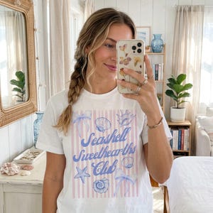 Könnte beinhalten: Weißes T-Shirt mit einem Muscheldesign und dem Text "Seashell Sweethearts Club" in blauer Schrift. Das Design umfasst Muscheln, Schleifen und einen Seestern auf einem rosa gestreiften Hintergrund. Das Shirt hat kurze Ärmel.