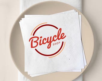 May include: A stack of white paper napkins with a textured pattern, featuring the red script text "Bicycle Shop" inside a red and orange circle. The napkins are on a beige plate.