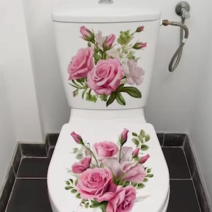 Può includere: Un WC bianco con un adesivo floreale decorativo sia sul serbatoio che sul coperchio. Il motivo mostra un gruppo di rose rosa, boccioli e foglie verdi. Un tubo di alimentazione dell'acqua in metallo argentato è fissato al lato del serbatoio.