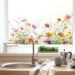 May include: A kitchen window with a floral design featuring colorful flowers in shades of pink, orange, and yellow. A white container labeled "COFFEE SUGAR" sits on the windowsill. A stainless steel faucet and sink are in the foreground.
