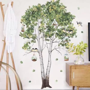 Könnte beinhalten: Ein Wandaufkleber mit einem großen Baum mit grünen Blättern und weißer Rinde. An den Ästen des Baumes hängen mehrere gläserne Terrarien mit Pflanzen, und kleine Vögel sitzen auf den Zweigen. Ein kleiner Vogel fliegt rechts davon.