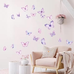 May include: A collection of watercolor butterfly wall decals in shades of purple, pink, and blue. The butterflies are scattered across a light pink wall, creating a whimsical and decorative accent. A pink armchair and side table are also visible.