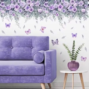 Puede incluir: Un sofá de terciopelo lavanda con un cojín a juego se coloca frente a una pared blanca adornada con calcomanías de rosas y mariposas moradas. Una pequeña mesa auxiliar blanca sostiene un jarrón morado con vegetación.