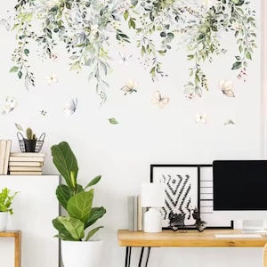 Könnte beinhalten: Eine weiße Wand, verziert mit einem Blumen- und Schmetterlingsaufkleber. Das Design zeigt kaskadierende grüne und weiße Blätter, zarte Schmetterlinge und kleine Blüten. Im Vordergrund befindet sich ein Schreibtisch mit Computer und Dekoration.
