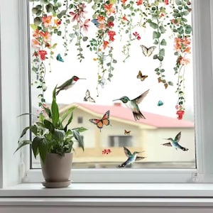 Op de afbeelding: Een raamsticker met kolibries, vlinders en hangende wijnstokken met rode, oranje en roze bloemen. Het ontwerp creëert een door de natuur geïnspireerde scène, met een potplant op de voorgrond.