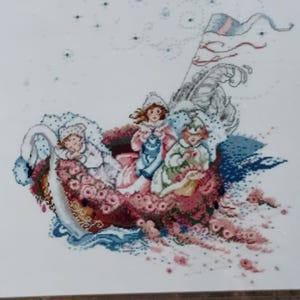 May include: Cross-stitch artwork depicting three figures in a floral boat with a swan-like design. The boat is filled with pink roses and blue accents. The artwork is framed in a dark wooden frame.