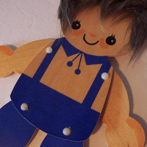 May include: A wooden toy doll with brown hair, blue overalls, and a painted smile. The doll has movable arms and legs.