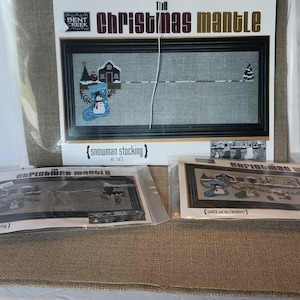 May include: A cross-stitch kit titled "The Christmas Mantle" featuring a snowman stocking design. The kit includes a framed piece with a gray background and a black frame, along with two additional kits.