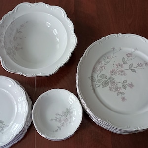 May include: A collection of white porcelain dinnerware with a floral design. The set includes various sizes of plates and bowls, all with a scalloped edge and a delicate pink and green floral pattern. The plates are stacked, and the bowls are arranged around them.