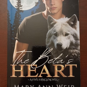 May include: Book cover with a black background and gold lettering. The title is "The Beta's Heart" with the subtitle "A Five Fangs Novel". The author's name is "Mary Ann Weir". The cover features a young man with blond hair and a white wolf.